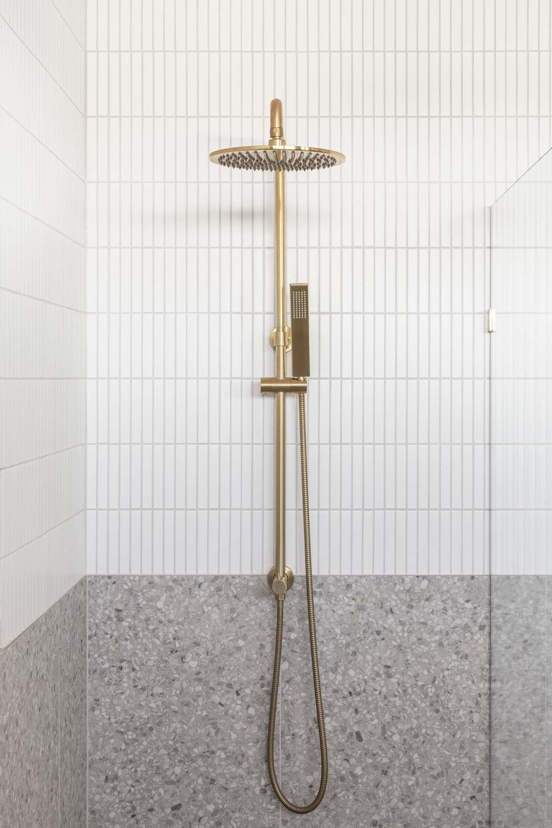 A-brushed-brass-dual-shower-set-in-a-frameless-shower-with-a-half-wall-of-grey-terrazzo-tiles-and-white-finger-tiles