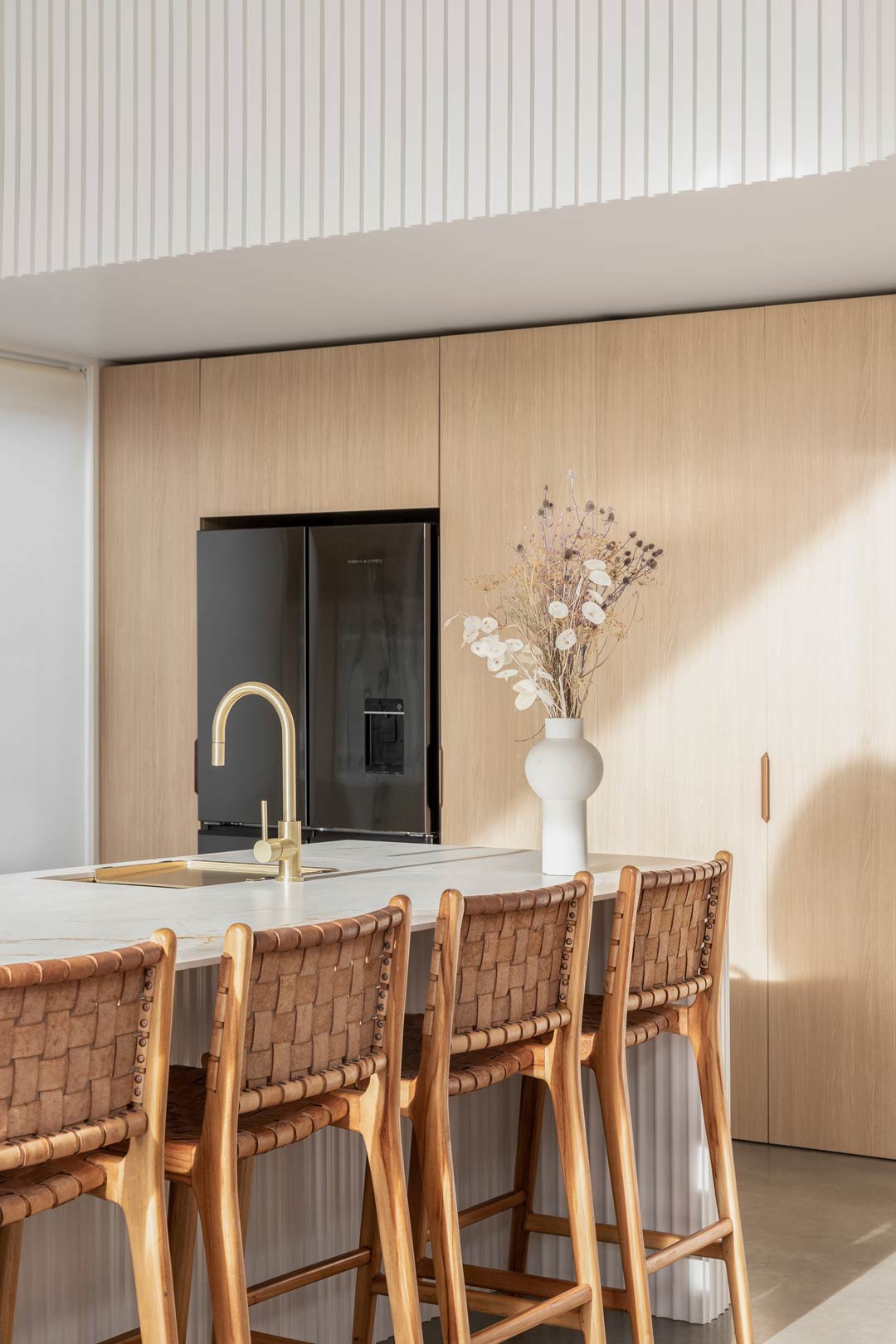 A-warm-open-kitchen-in-a-barn-style-house-with-large-island-bench-brushed-brass-tapware-timber-cabinetry-and-woven-leather-bar-stools