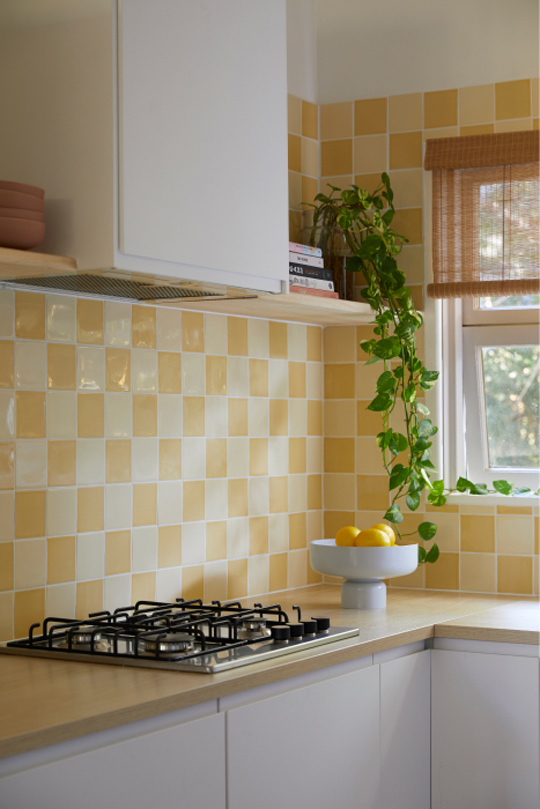 Two-toned-square-yellow-splashback-tiles-a-white-kitchen-and-a-timber-bench-top-with-a-timber-shelf-with-draping-plant