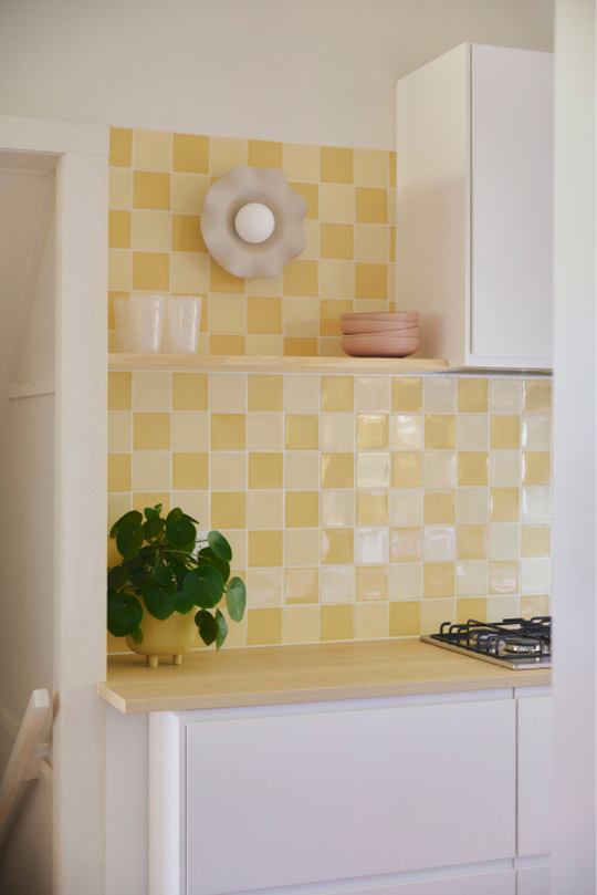 Two-toned-square-yellow-splashback-tiles-a-white-kitchen-and-a-timber-bench-top-with-a-timber-shelf